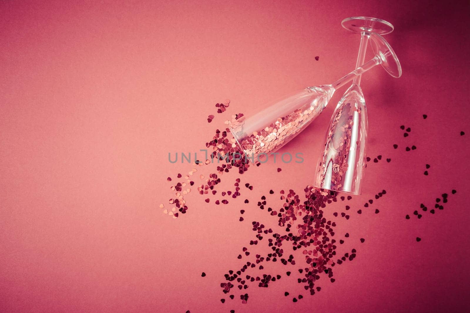 Two champagne glasses with splash of red heart shaped confetti on red background. Overhead view, copy space. Valentine's Day concept