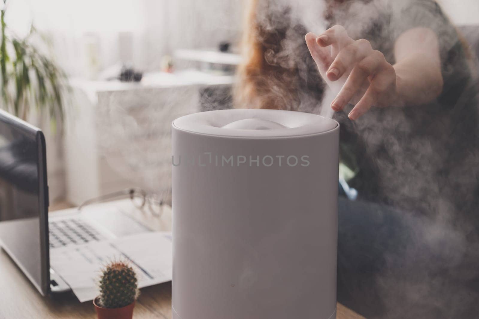 Woman freelancer uses a household humidifier in the workplace to maintain relative humidity and microclimate in the workplace of the home office with a laptop and documents.