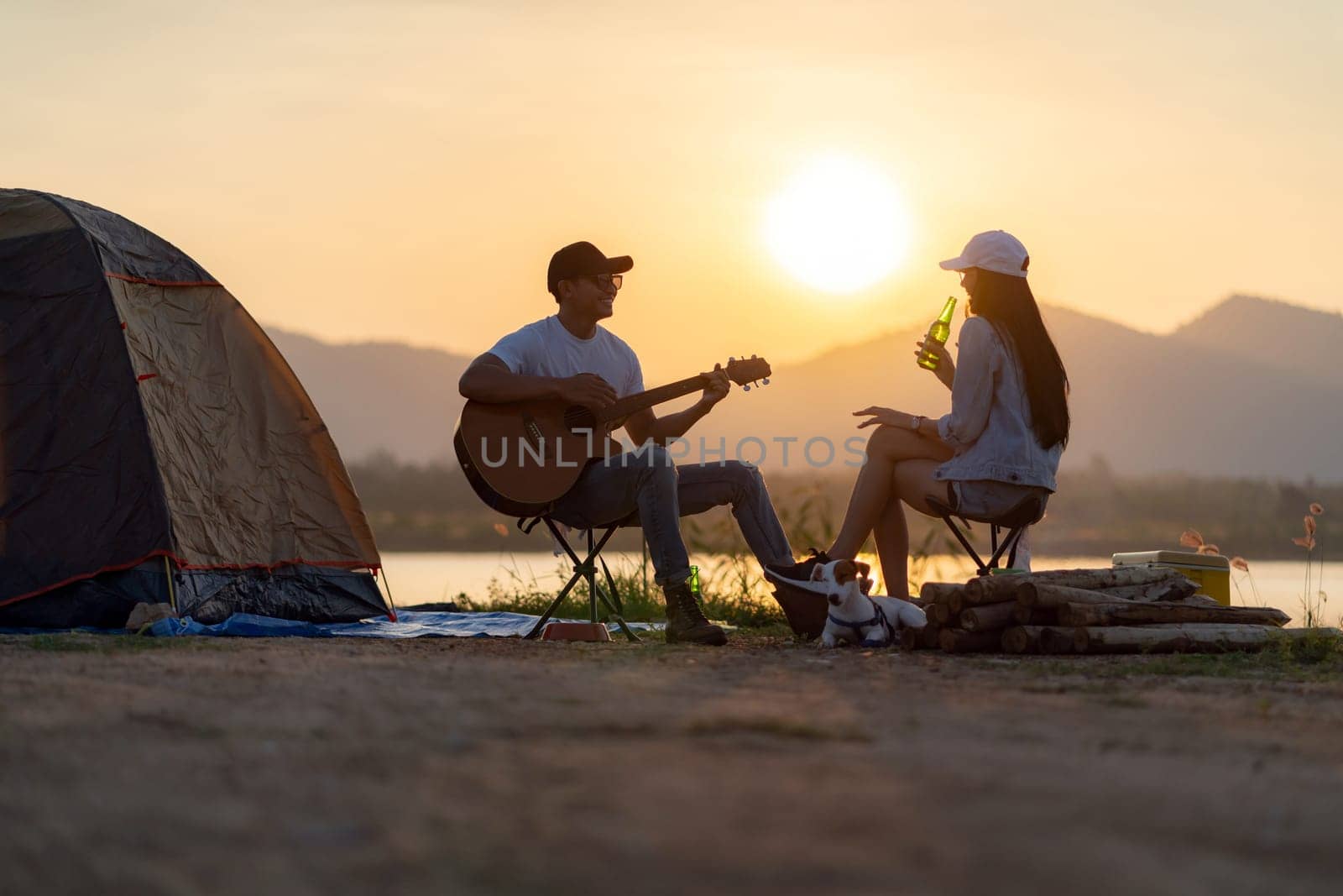 Young adult Asian couple playing guitar and drinking beer beside their tent campsite while sunset. Camping trip with dog outdoor activity campsite concept.