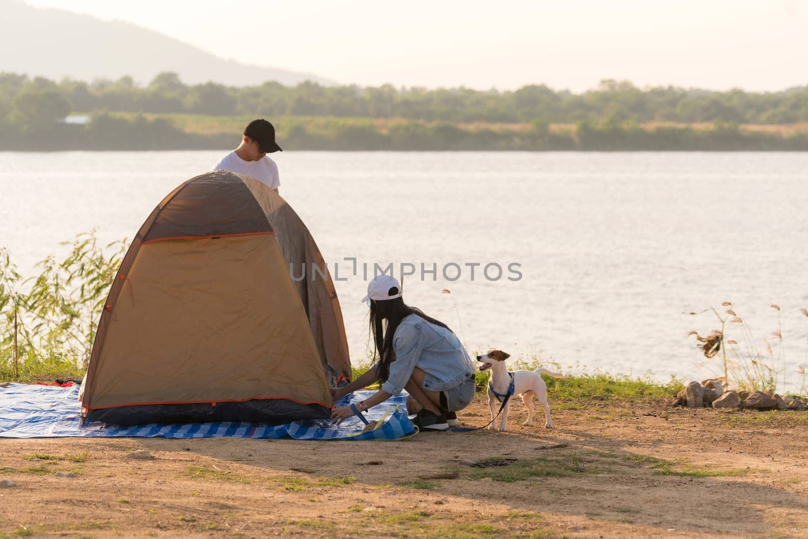 Young adult Asian couple pitch and set up tent for camping around the lake. Camping trip with dog outdoor activity campsite concept.