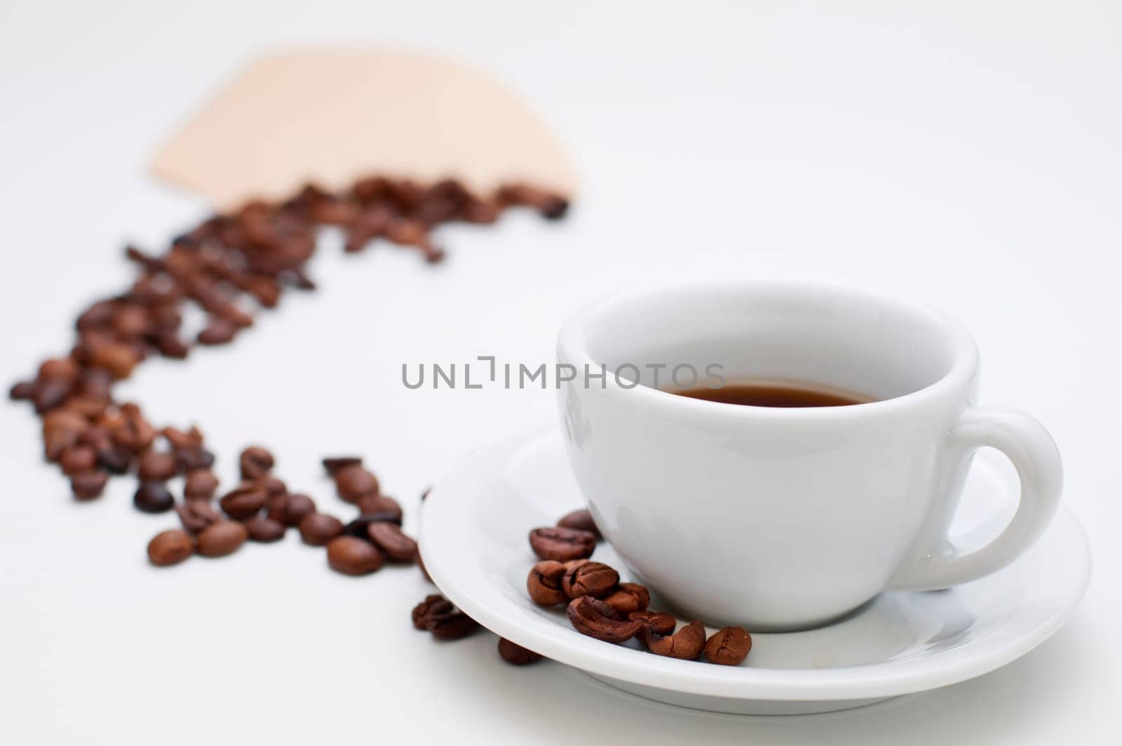 White half-filled cappuccino cup with saucer and individual roasted coffee beans in the sharp foreground and coffee beans arranged in an arch towards a filter bag in the blurred background on a white background.