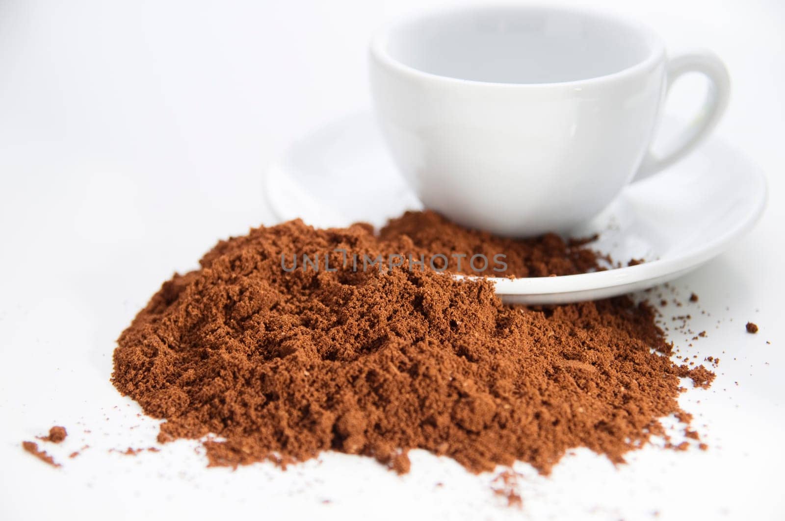 White cup and saucer on a white background with coffee powder scattered in front of it, which extends to the saucer.