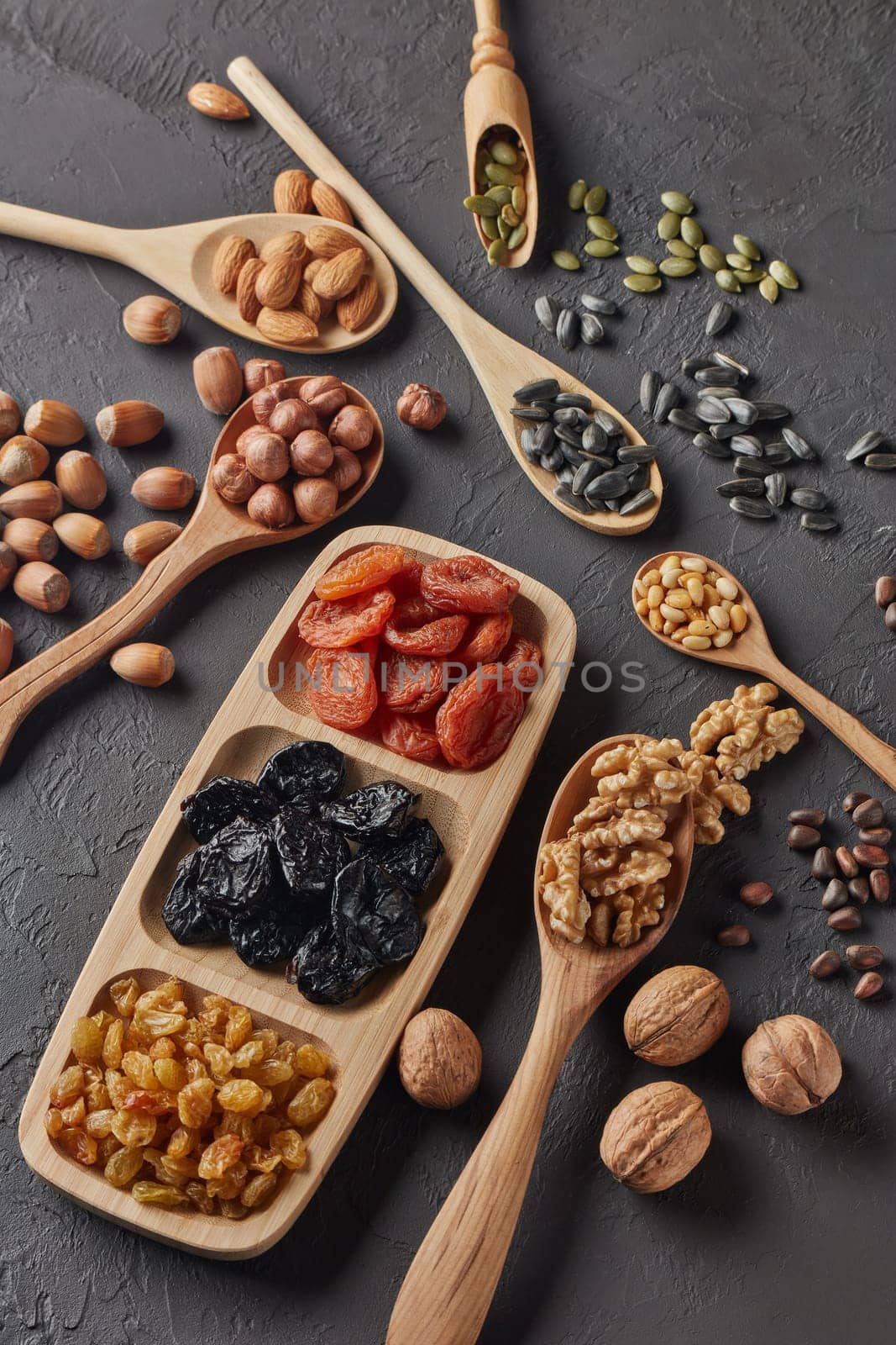Various varieties of nuts, seeds and dried fruits lying in wooden spoones and dish on black slate background. Top view. Healthy food. Vegetarian nutrition