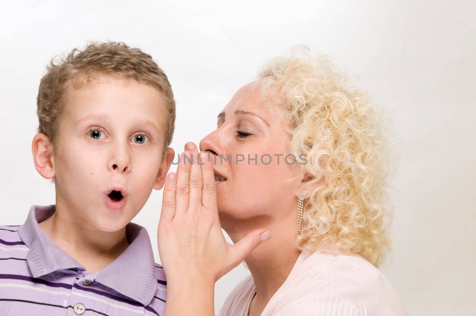Head-and-shoulder view blonde mother whispers sideways into her 7-year-old son's ear with her hand held against a white background