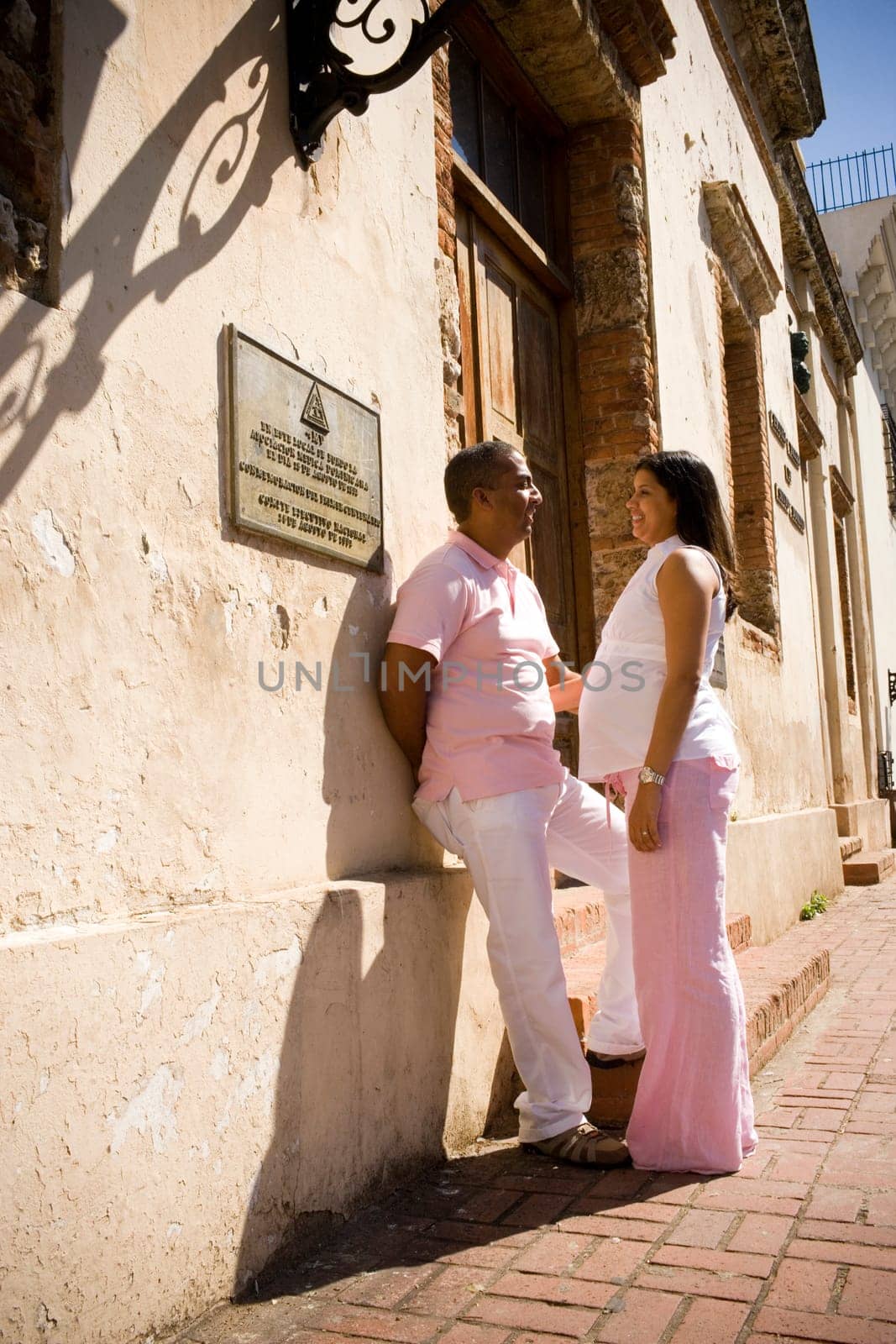 Side profile of a mid adult man leaning against a wall with a pregnant young woman standing in front of him