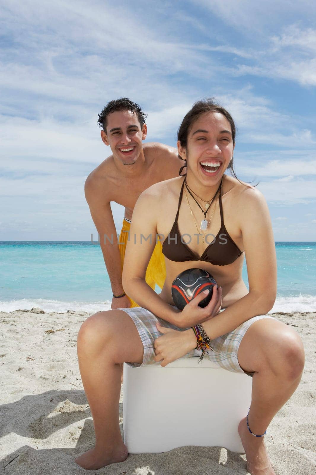 Portrait of a young woman sitting on an ice box on the beach with a young man standing behind her
