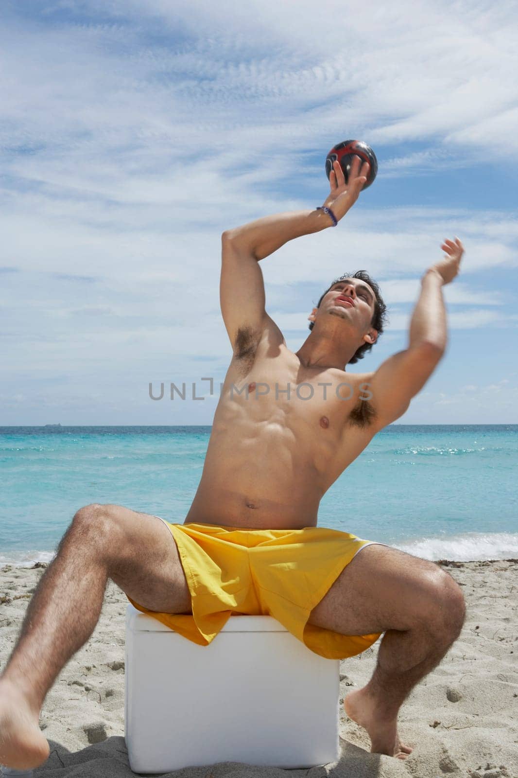 Young man sitting on an ice box on the beach and playing with a soccer ball