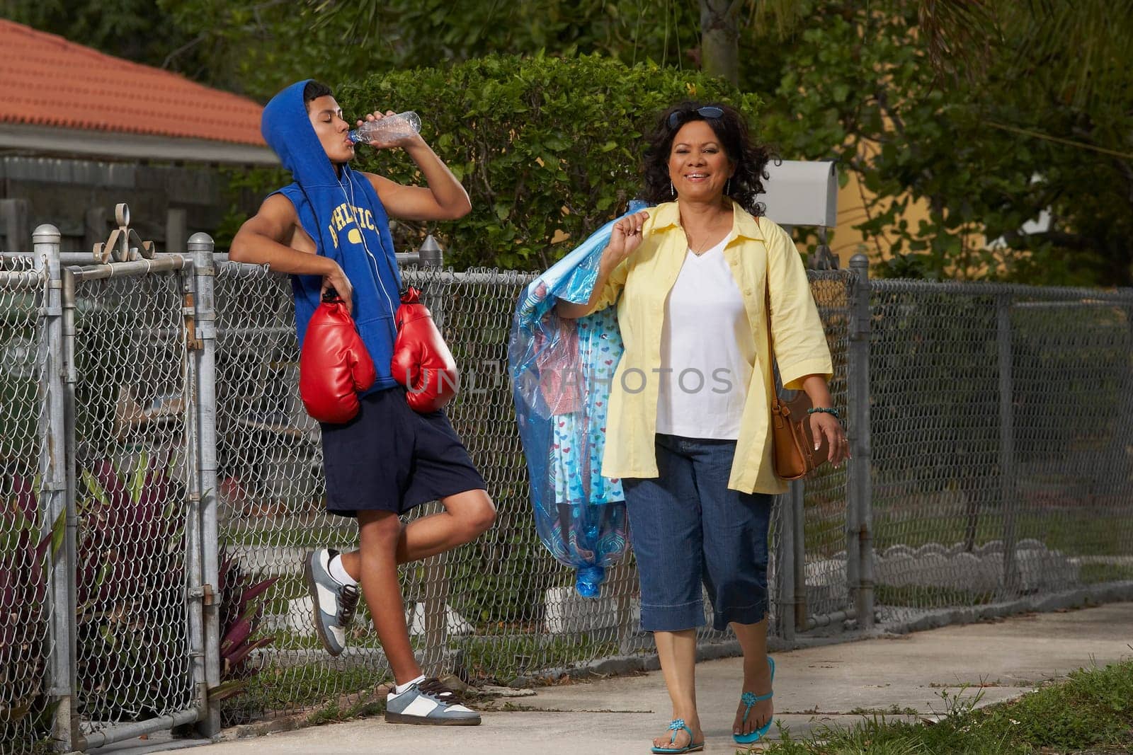 Mature woman walking with a teenage boy leaning on a chain-link fence and drinking water beside her