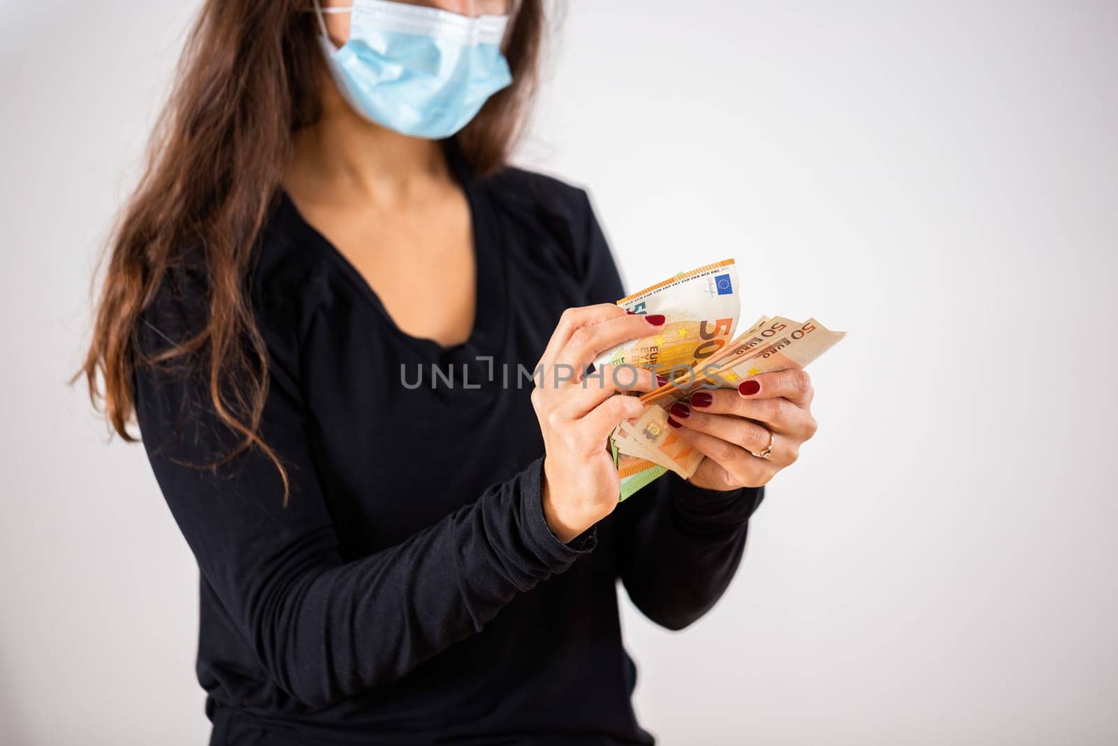Brown haired woman holding money in her hands with face mask covering her mouth and nose. Girl counting euro banknotes with nominal values of fifty during coronavirus pandemic. Concept of poverty.