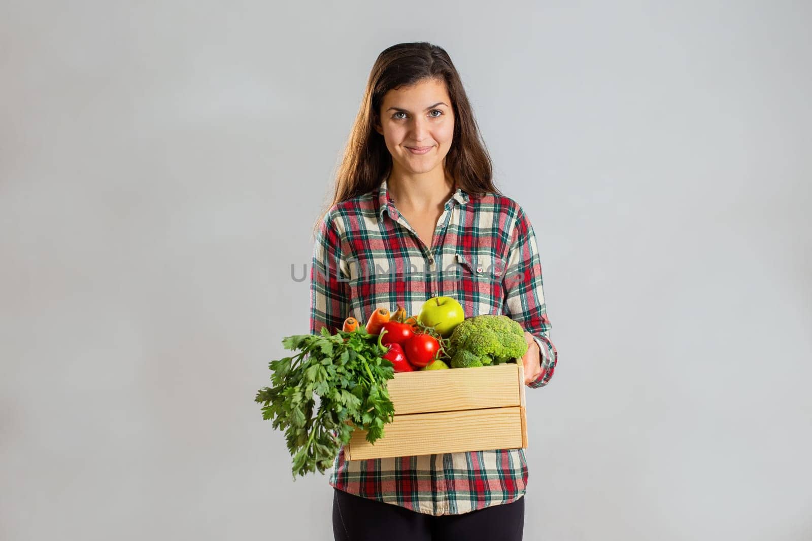 Young brunette holding a box full of fresh fruit and vegetable. Smiling woman with wooden crate filled with healthy ripe nutritious food. Positive lady in flannel shirt showing tasty organic plants.