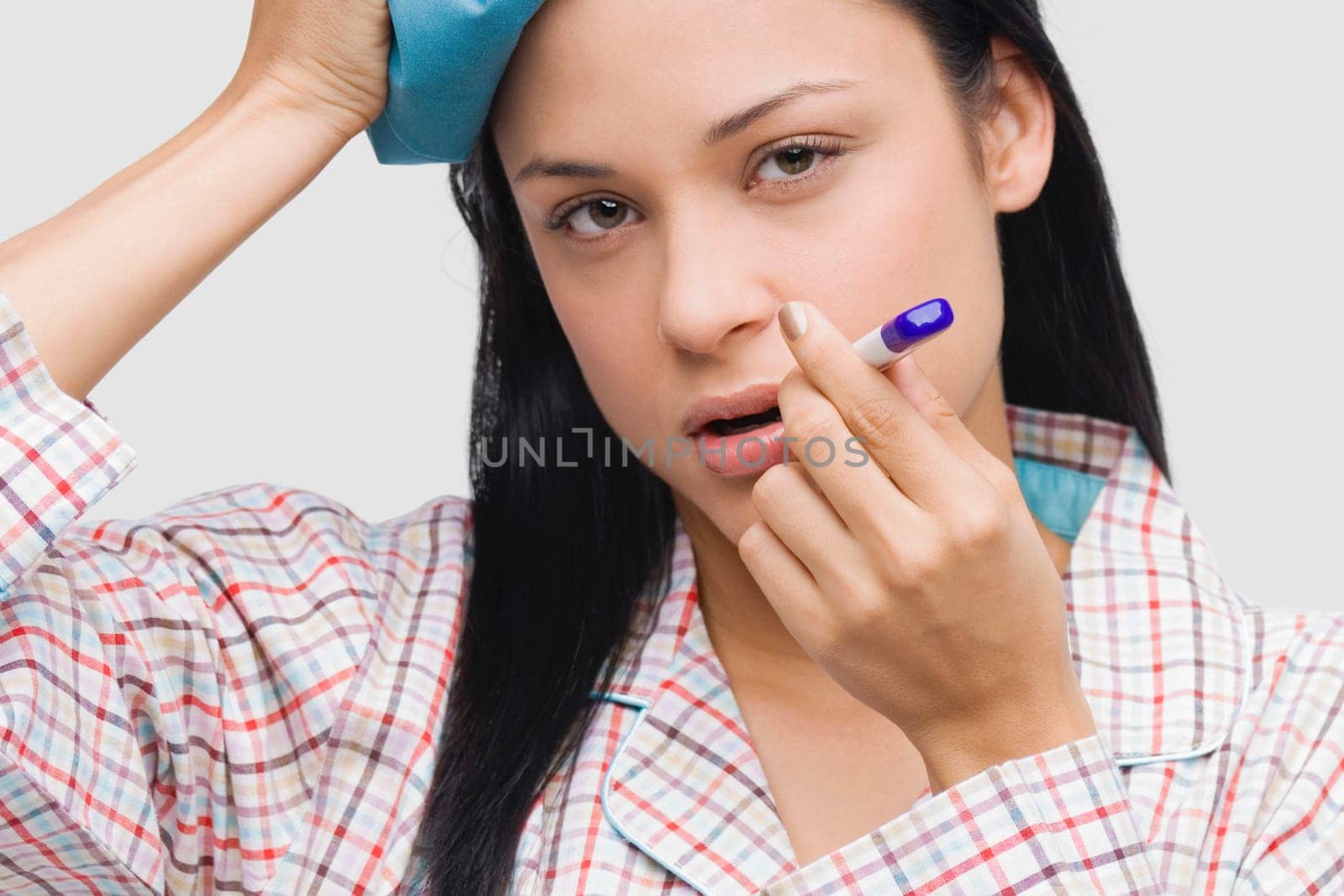 Portrait of a young woman measuring her temperature with a thermometer and holding an ice pack on her head