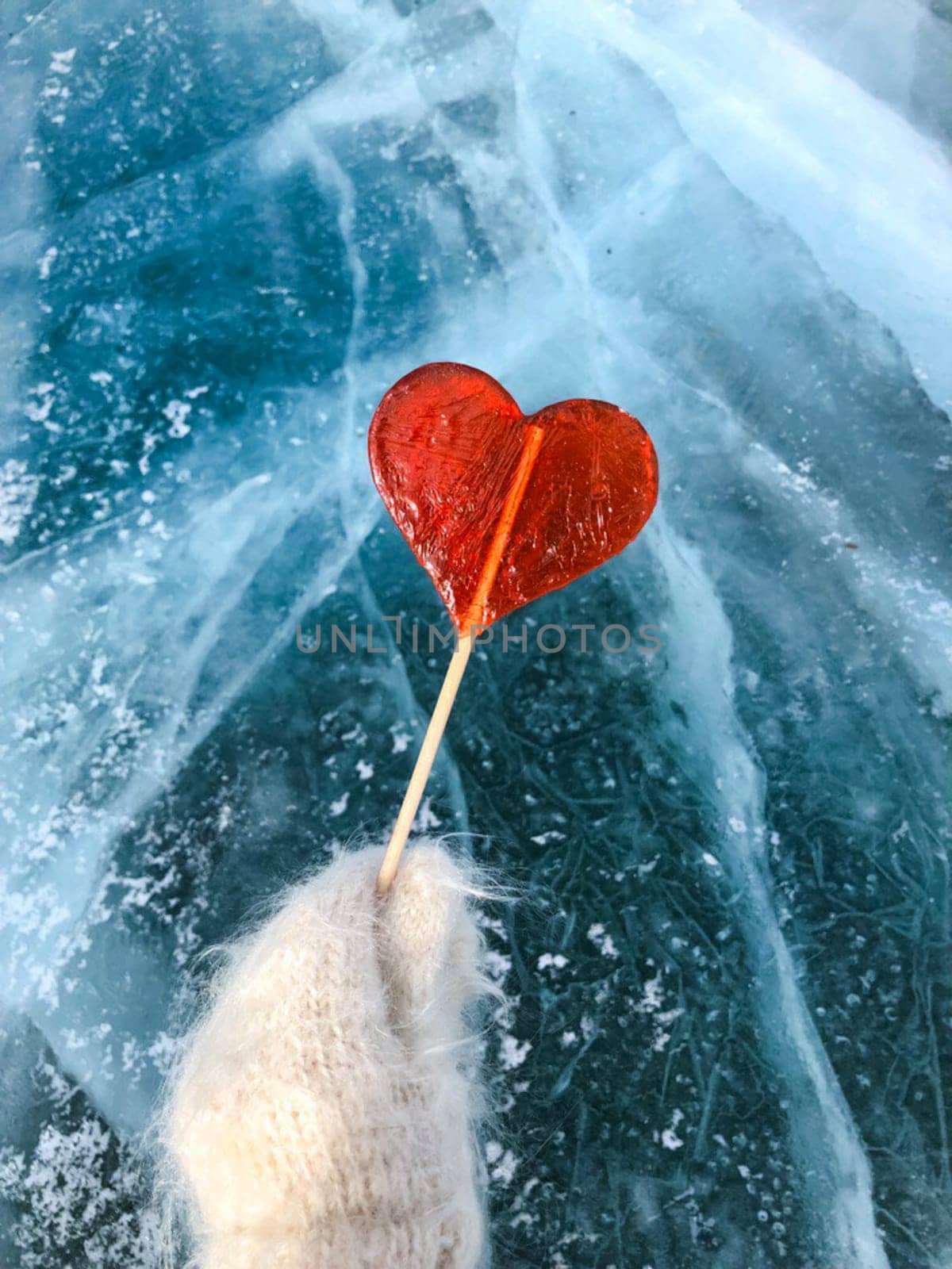 A red lollipop in the shape of a heart holds a hand in a mitten against the background of ice.