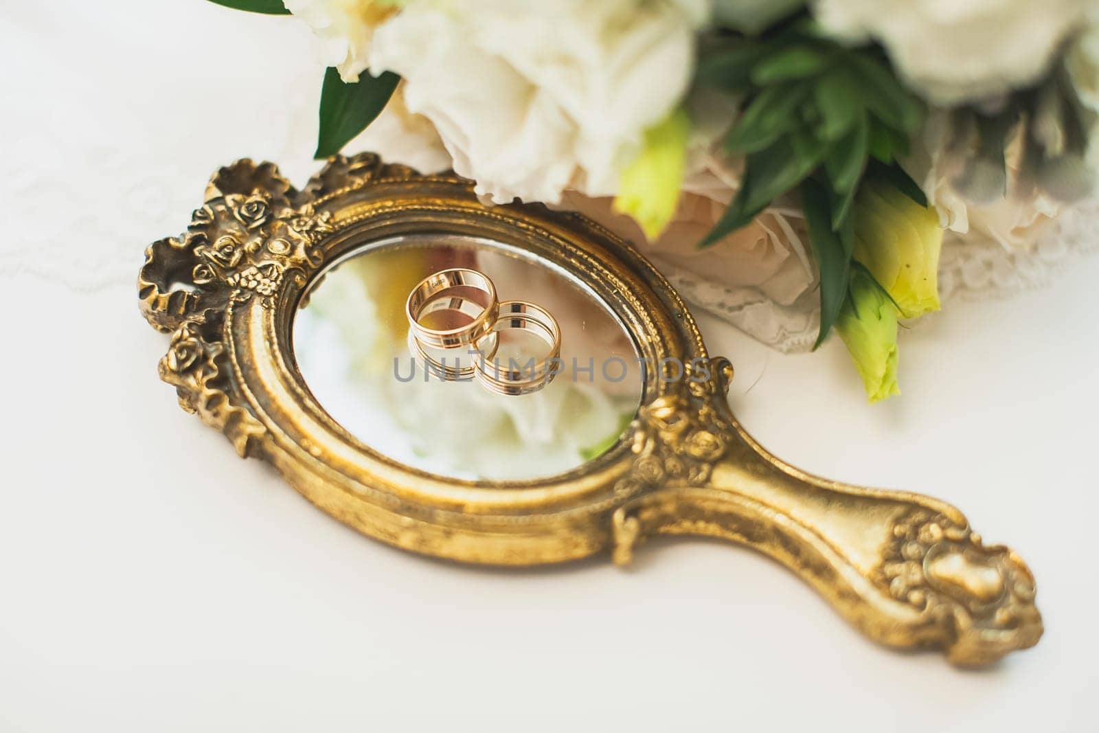 Wedding rings lie on a mirror with a gilded handle. Nearby is a bouquet of flowers.