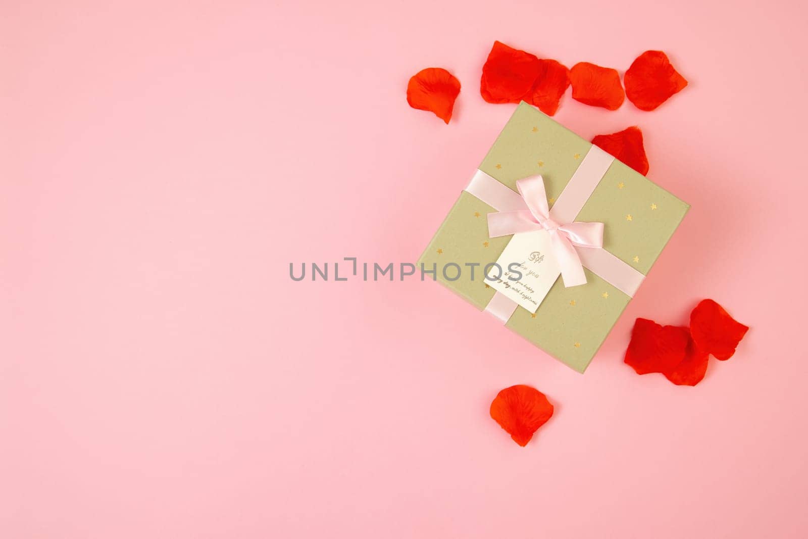 Pink background with present and rose leaves around. Golden box with ribbon from directly above. Gift lying on colorful table with copy space in flat lay composition.