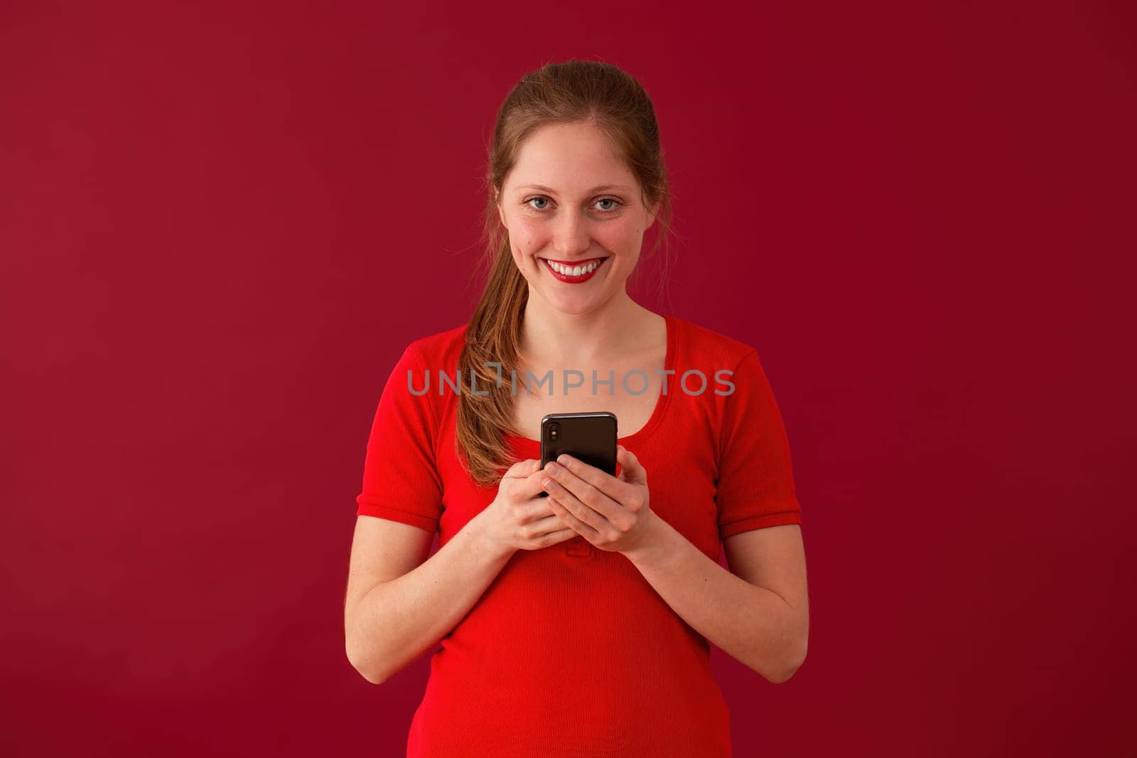 Blonde girl smiling with phone in hand on red background. Happy woman with lipstick texting on mobile on valentine. Positive female sending love message.
