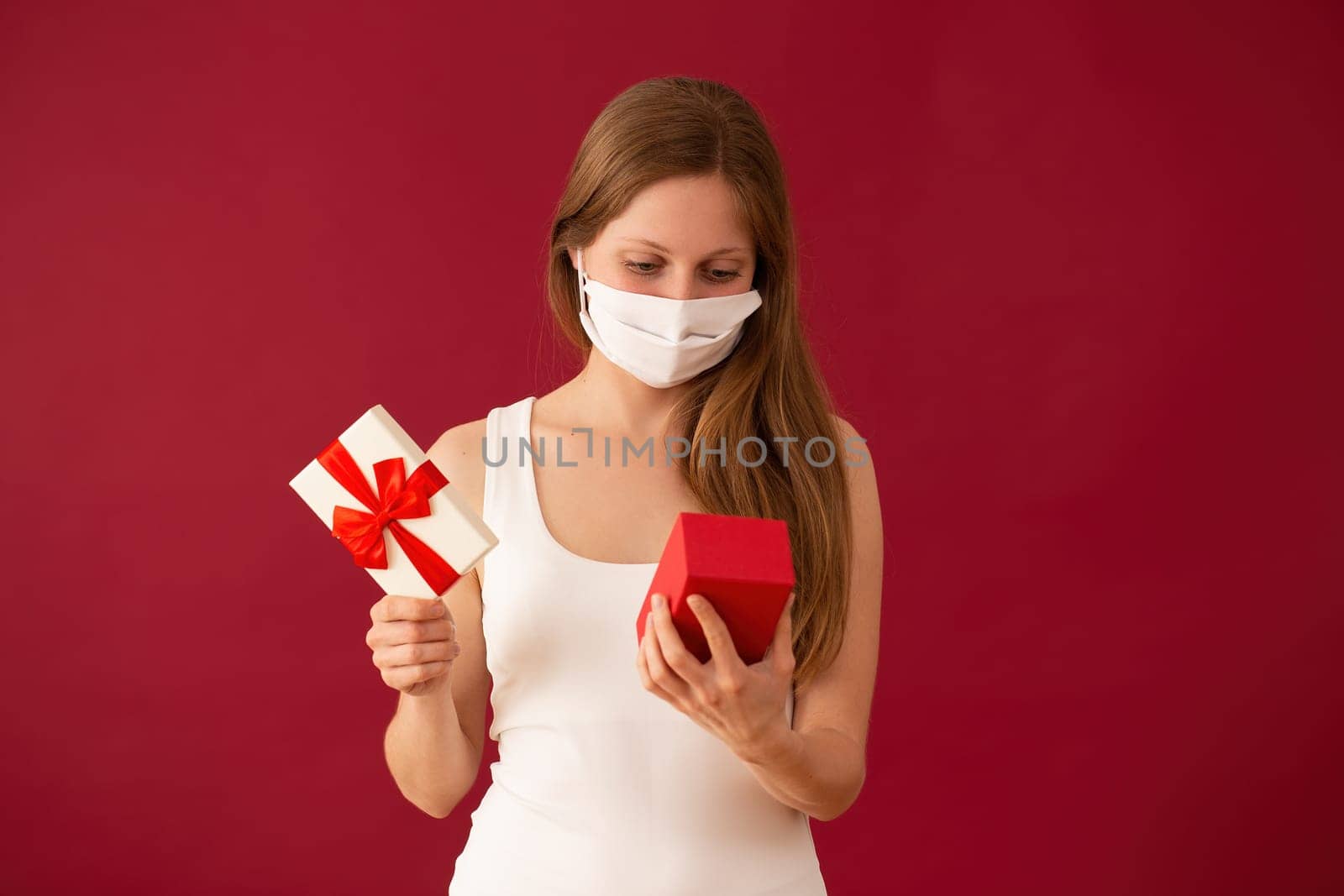 Girl with face mask opening present on valentines day. Blond woman holding box with ribbon on red background. Concept of love during coronavirus pandemic.