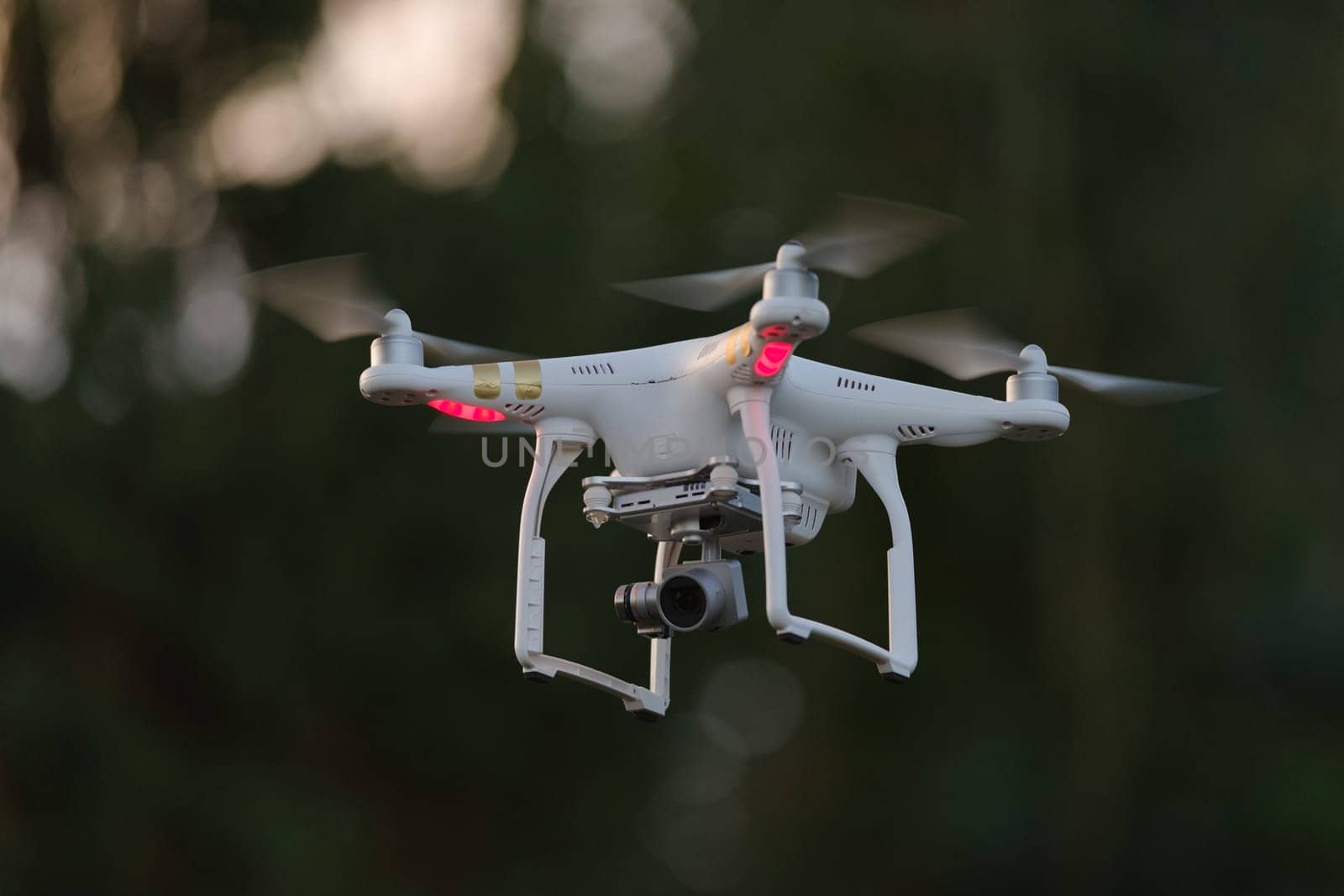 12 - White quad rotor drone with orientation lights hovering isolated from dark defocused background. Professional camera on gimbal beneath gives FPV
