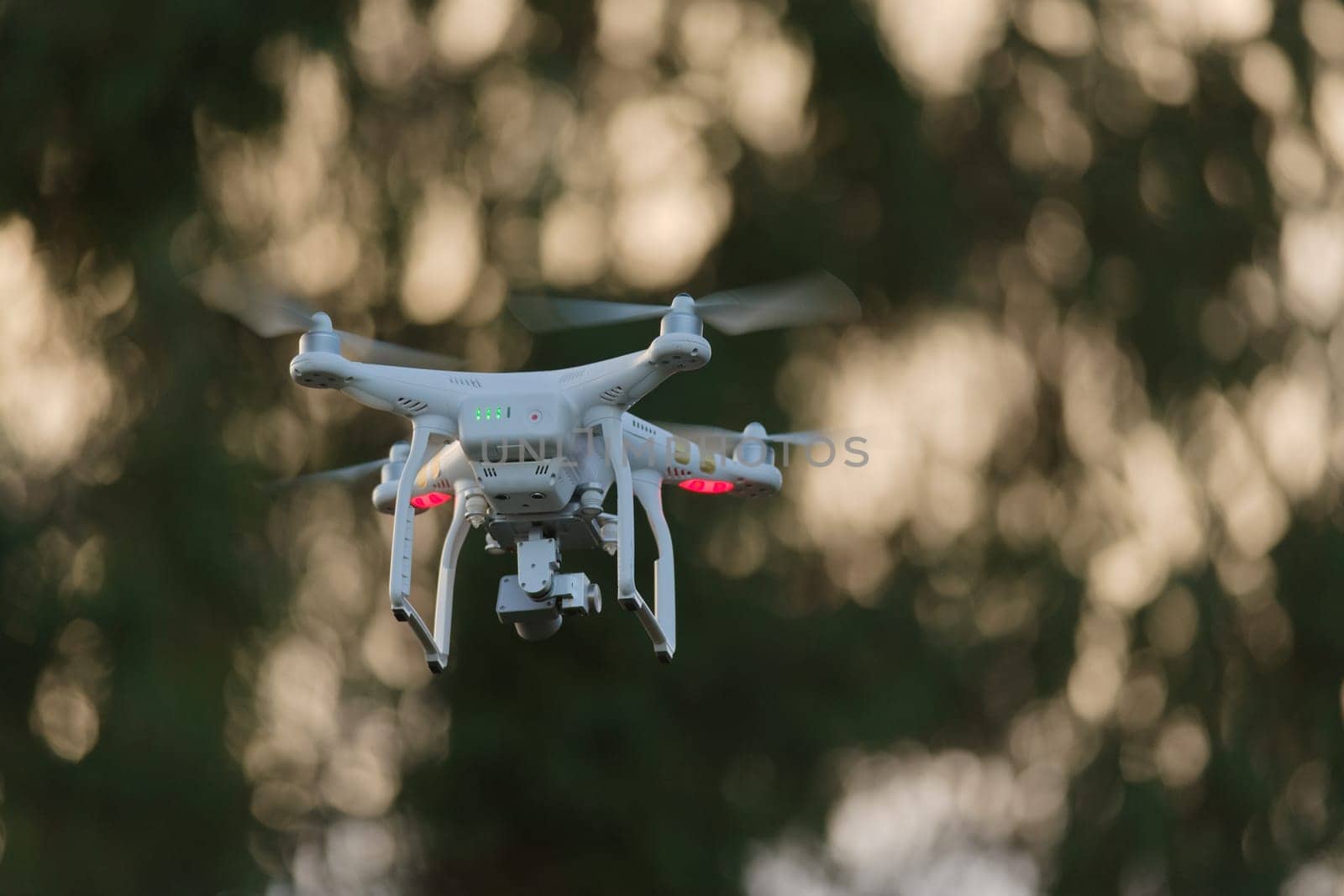 21 - Sunset bokeh background behind white quadcopter camera drone. Golden hour sun light creates dreamy smooth bokeh background shining through trees