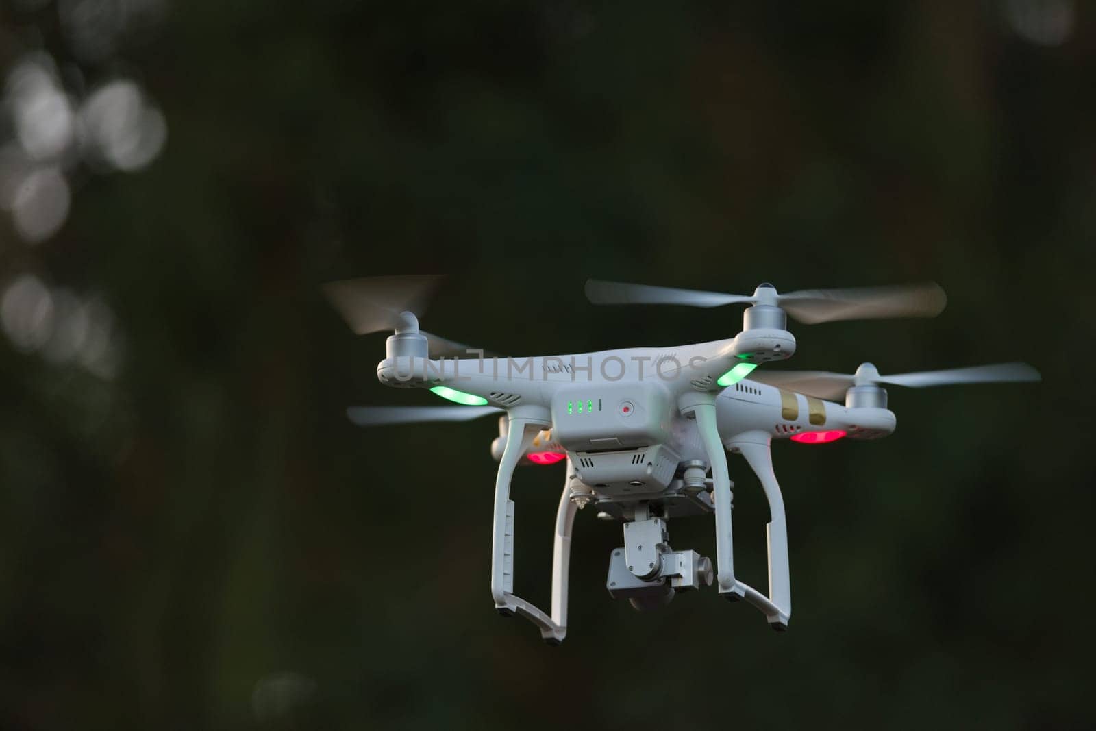 23 - Green and red orientation lights on white quadcopter camera drone as it flies towards dark out of focus trees