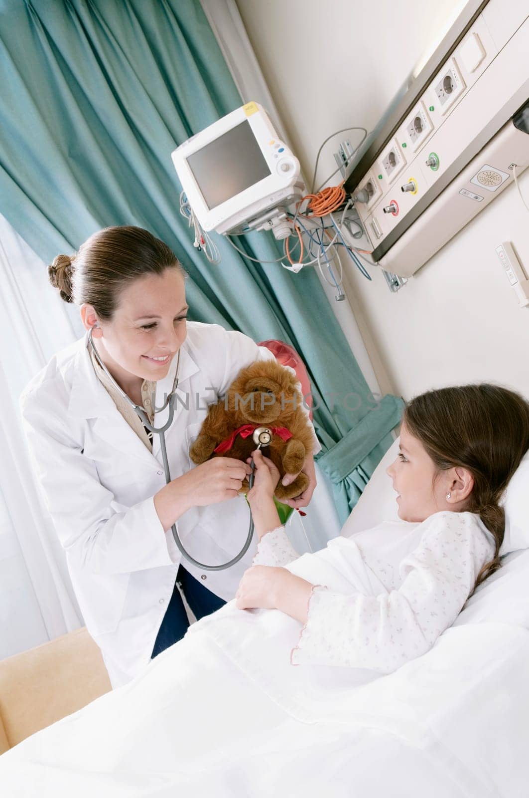Girl on a hospital bed with a female doctor examining a stuffed toy