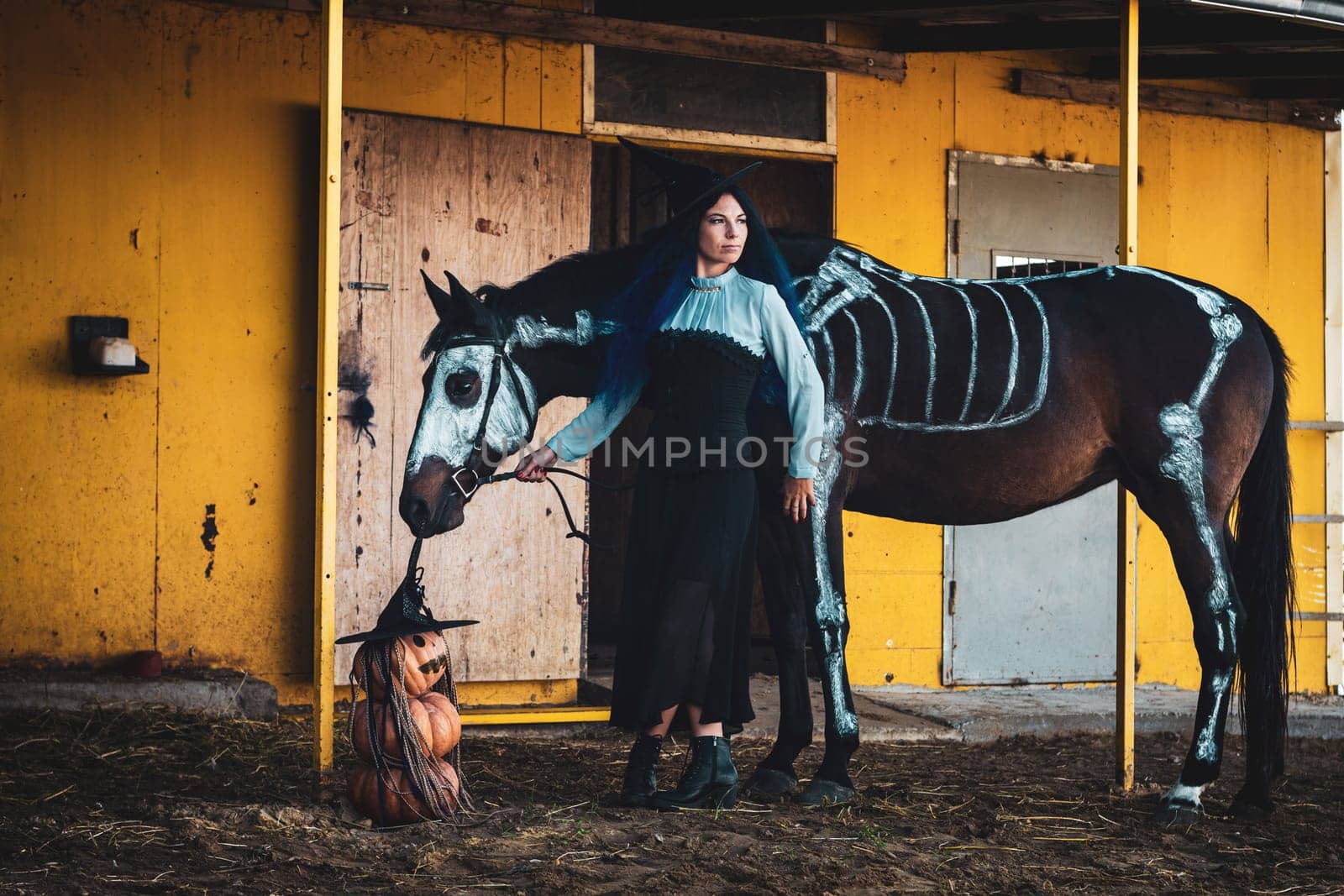 A girl dressed as a witch on a farm, a horse with a skeleton is drawn next to it, as well as three pumpkins in the shape of an evil man
