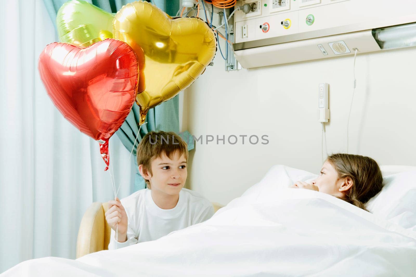 Girl on a hospital bed with her brother holding balloons