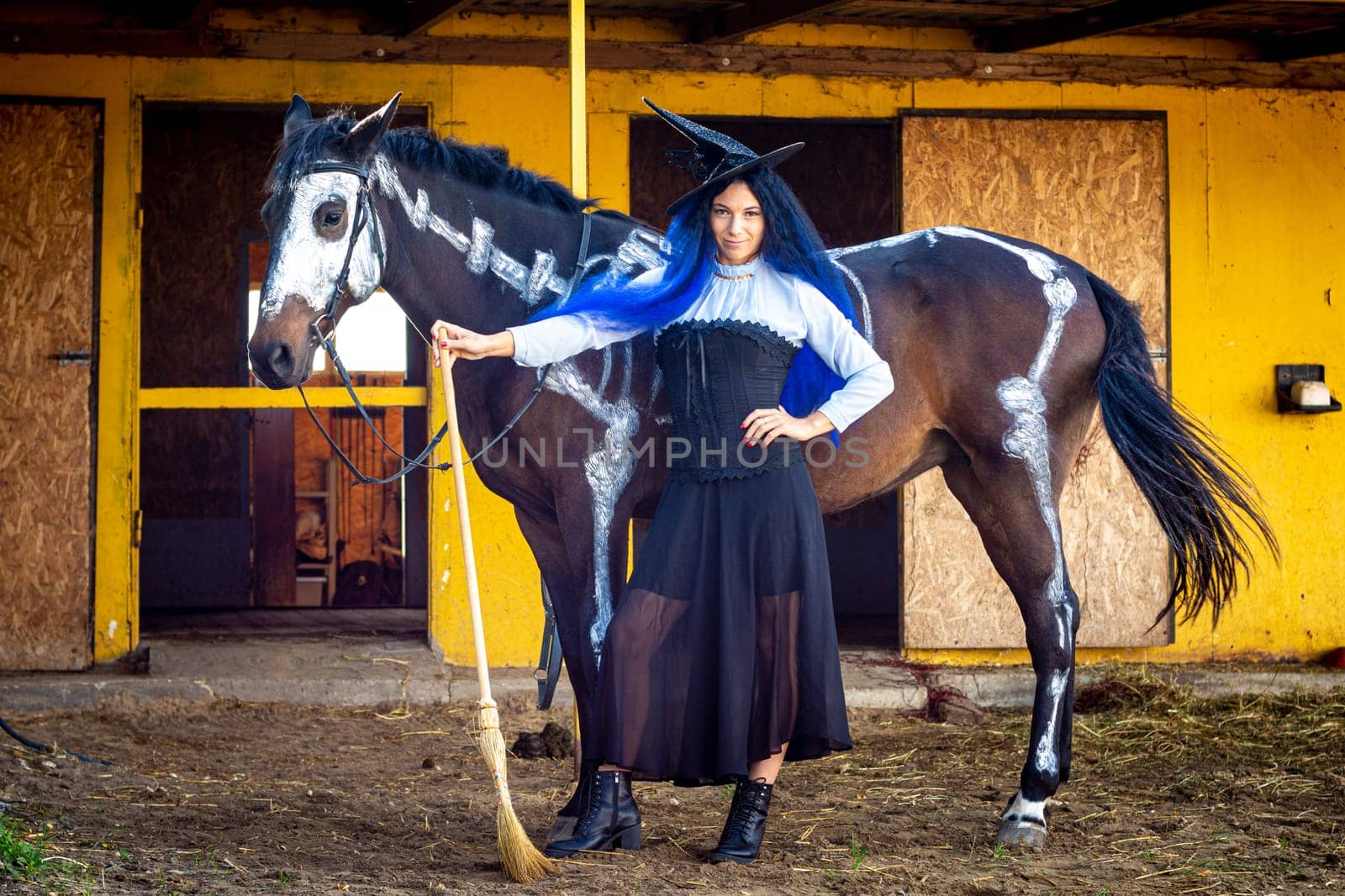 A girl dressed as a witch stands with a broom near a horse on which a skeleton is drawn
