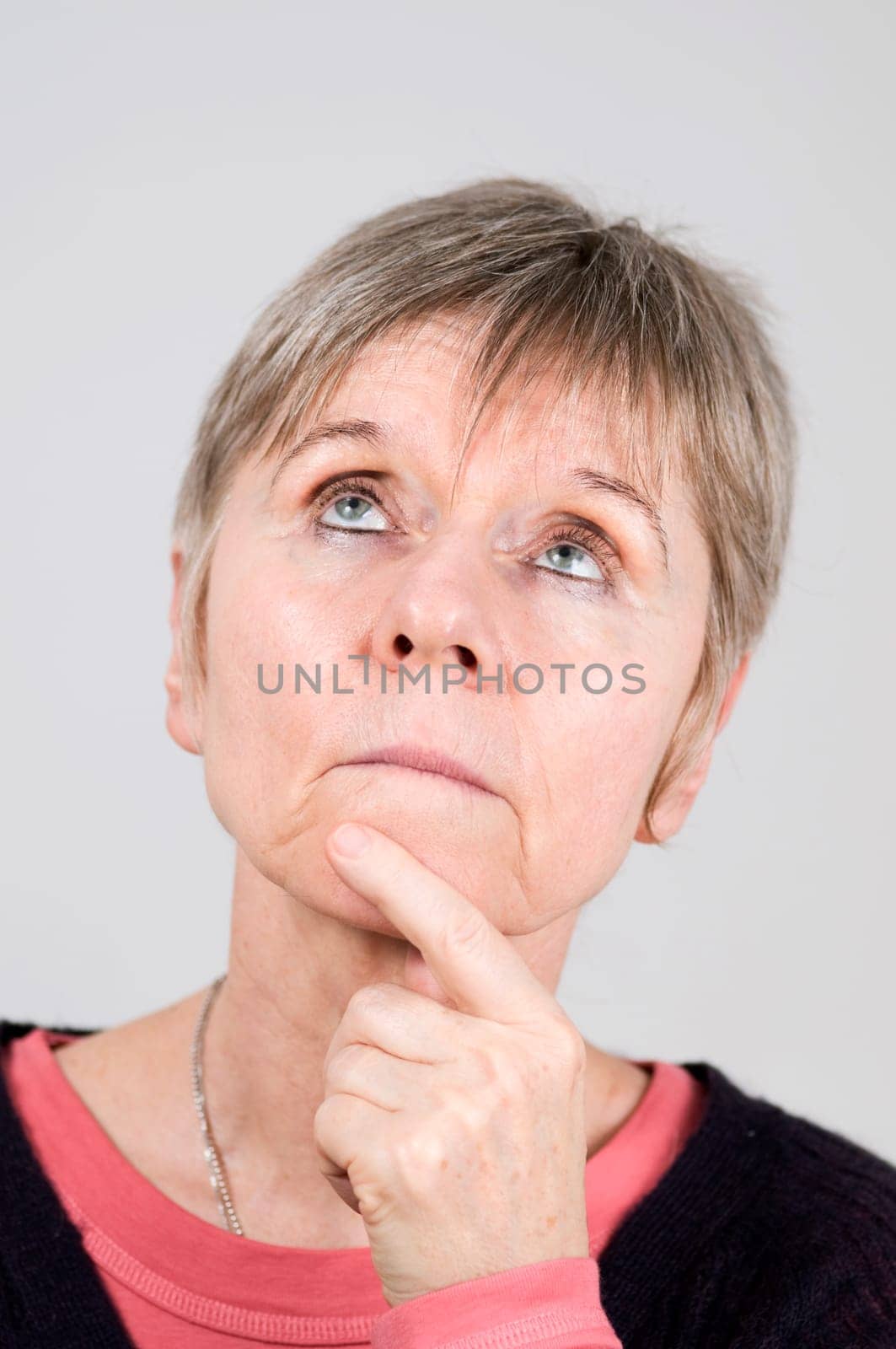 Head portrait of an older woman in front of a white background, pensive and serious looking upwards with her left index finger on her chin.