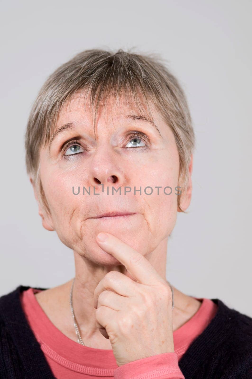 Head portrait of an older woman in front of a white background pensive and pressing lips, looking up with the left index finger on her chin.