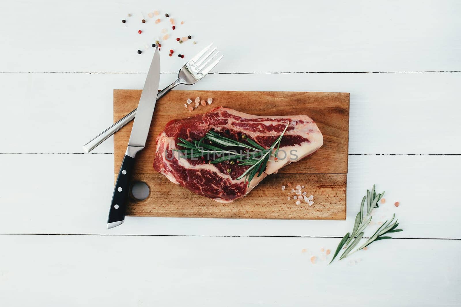 Raw ribeye steak on board, knife, fork, pepper, rosemary and pink Himalayan salt. High quality photo