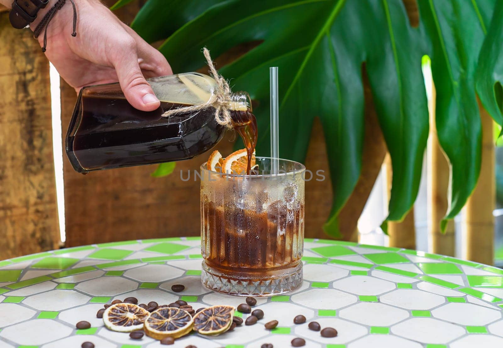 Hand pouring cold brew coffee in a bottle with transparent glass full of ice on green leaves background