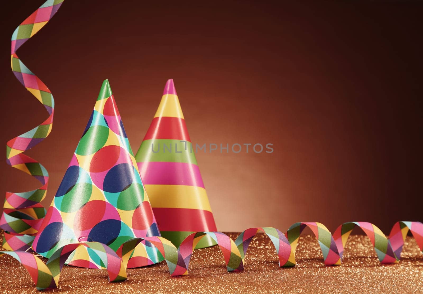 Close up Party Carnival Hats with Different Designs and Paper Streamers on Top of the Table, with Particles, in front Gradient Brown Background.