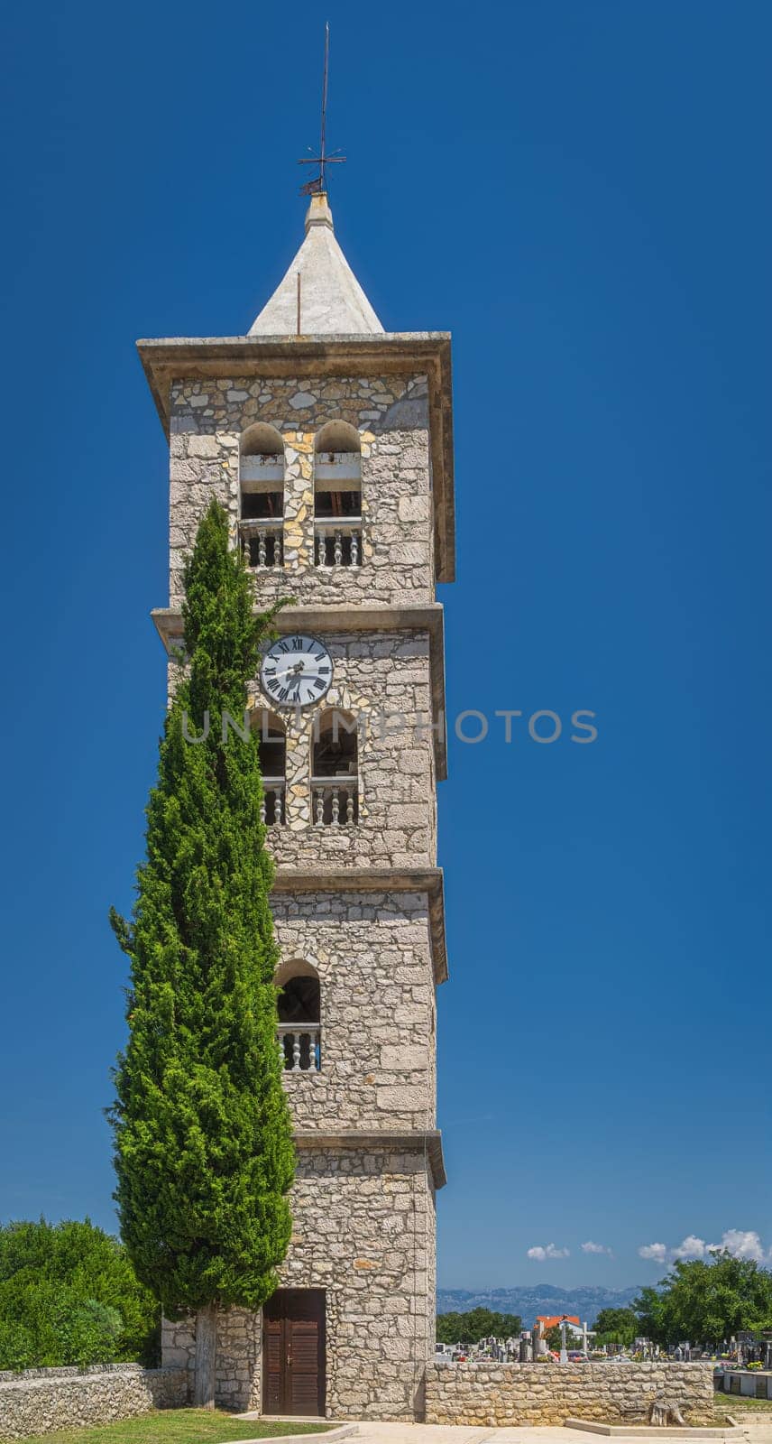 Bell and clock tower of a Church of the Nativity of the Blessed Virgin Mary in Zaton with graveyard and Dinaric Alps in a background, Croatia