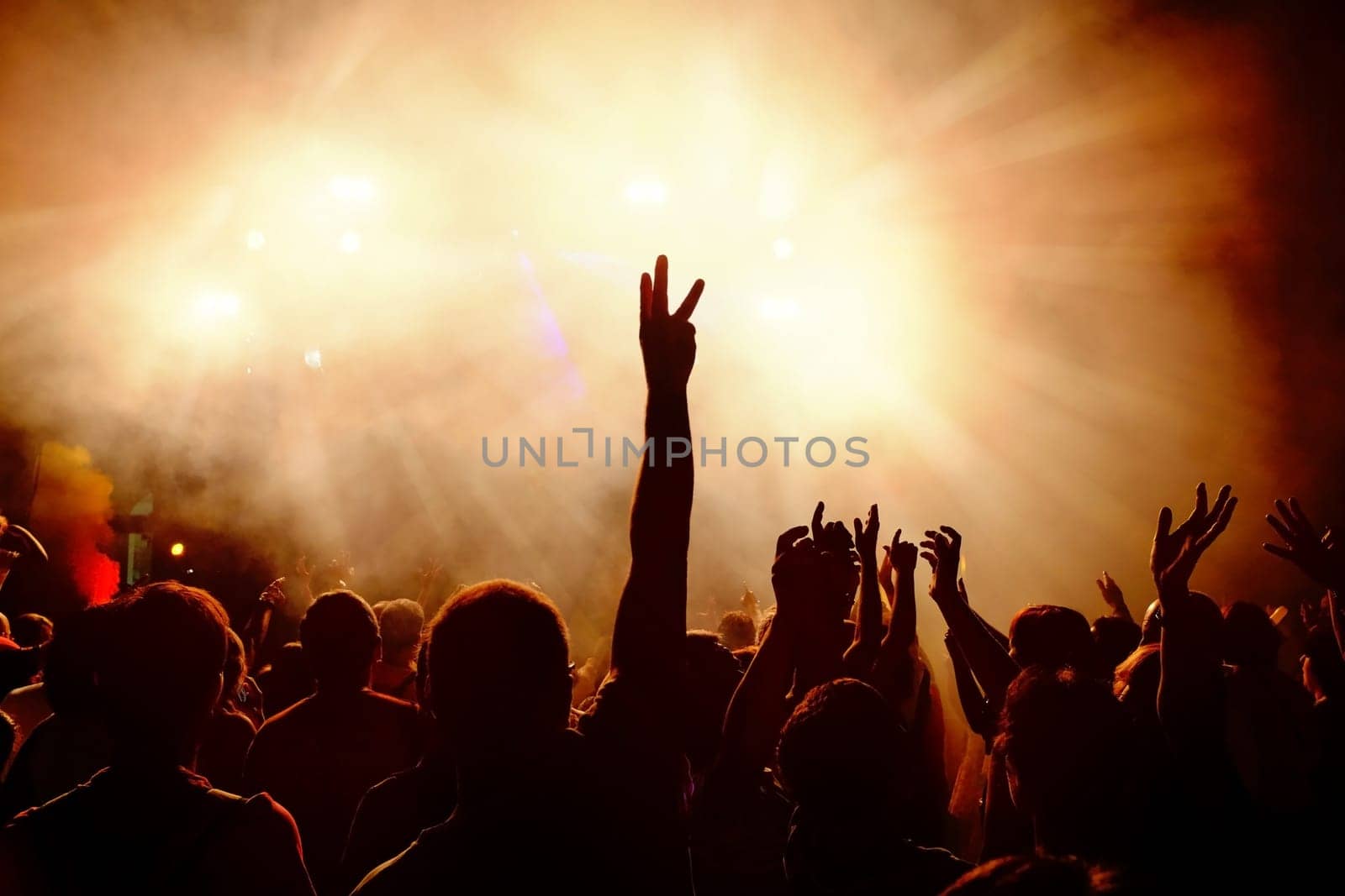 Silhouettes of festive crowd of young people dancing at music festival. Raising hands against smoky orange background with light beams.
