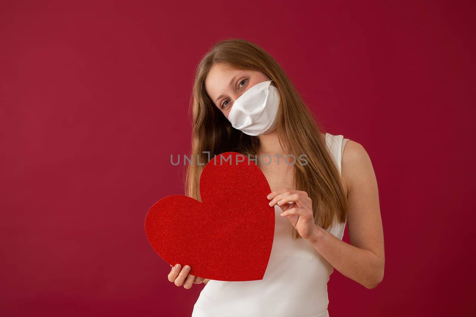 Cheerful woman with face mask showing red heart from front view. Optimistic young girl on valentine's day during coronavirus pandemic. Concept of love.