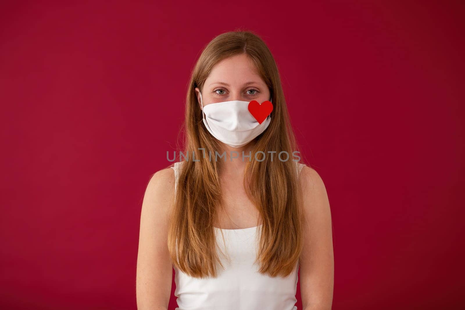 Woman wearing white cloth face mask and looking into camera from front view on red background. Concept of love during epidemy. Portrait of young person on valentine's day.