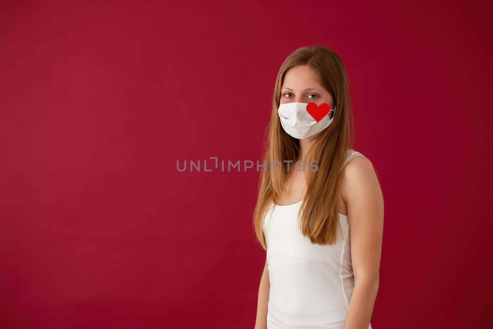 Solitary young Caucasian woman smiling under white face mask with red heart sign. Concept of celebrating valentine alone during coronavirus pandemic.