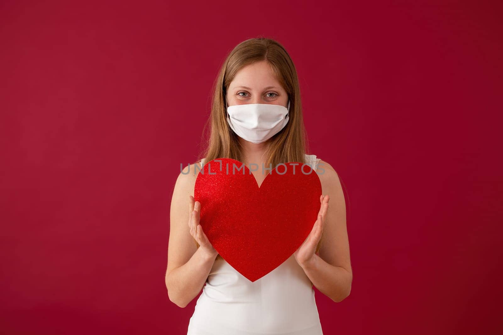 Smiling woman wearing face mask and holding red heart in hands. Concept of valentine during coronavirus pandemic. Cheerful blonde Caucasian girl from front view.