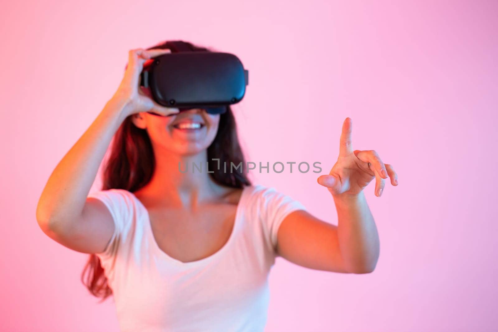 Brunette with headset pointing her finger in virtual reality. Smiling woman with digital glasses gaming in cyber space. Young female interacting in fictitious world.
