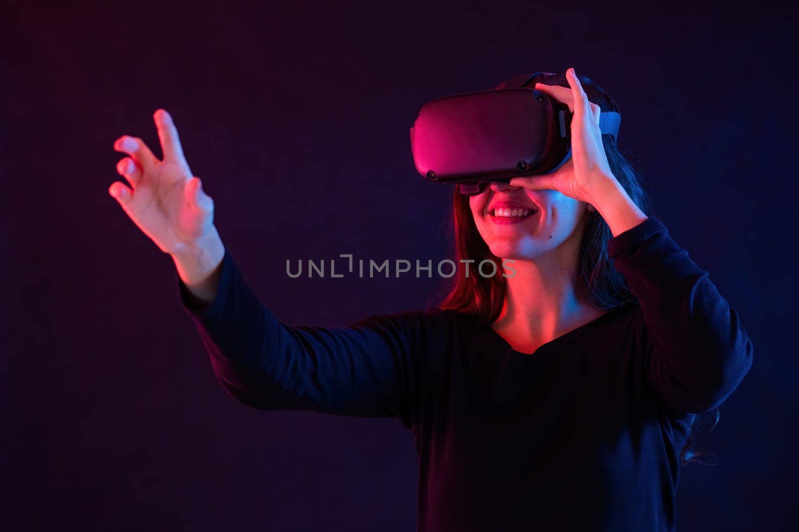 Smiling woman with headset interacting in virtual reality. Brunette using head equipment in dark space with red and blue light. Female touching fictitious thing.