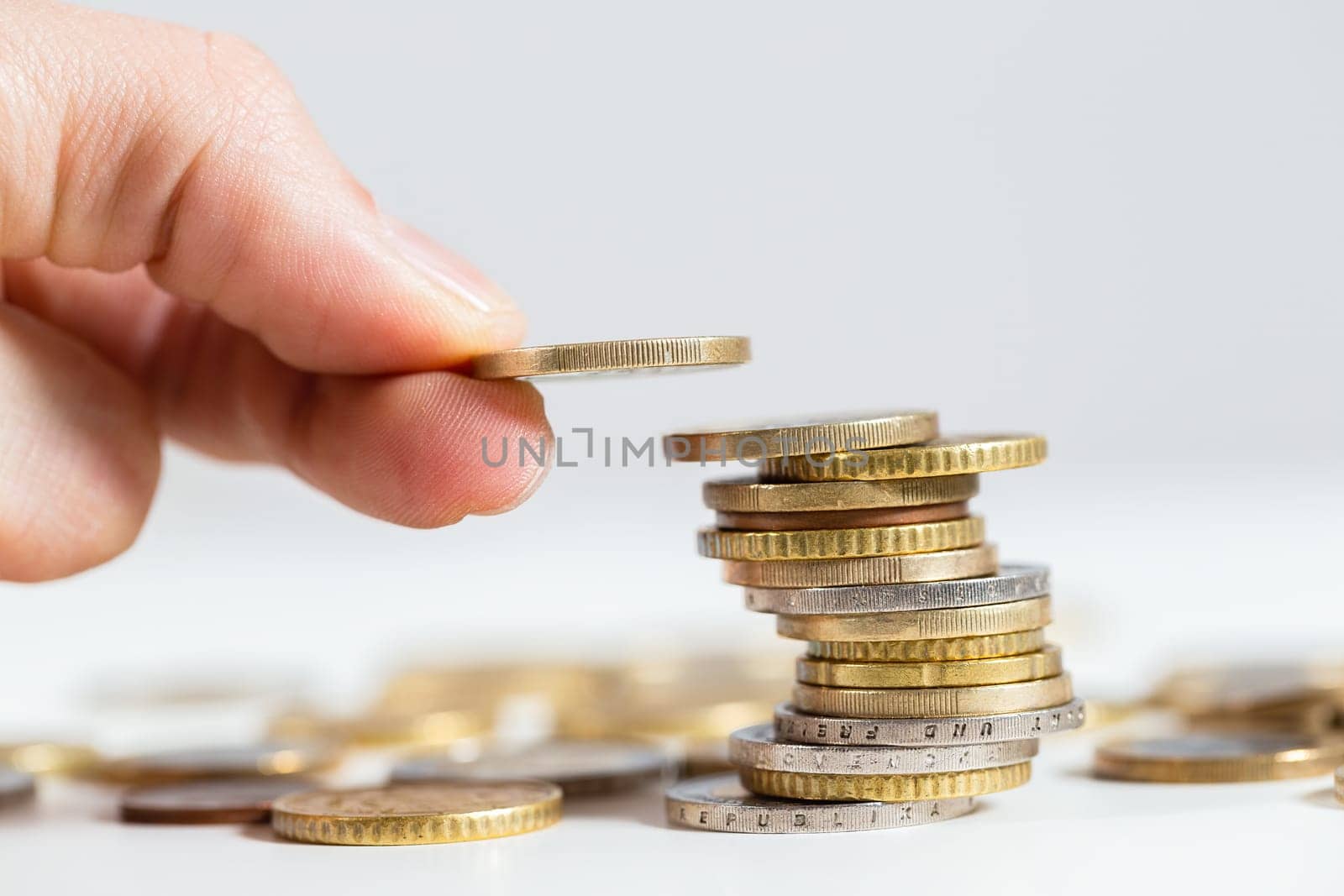 Holding coin in fingers over the stack of money from close up. Adding one euro on pile of metalic cash. Concept of saving salary for better profit.