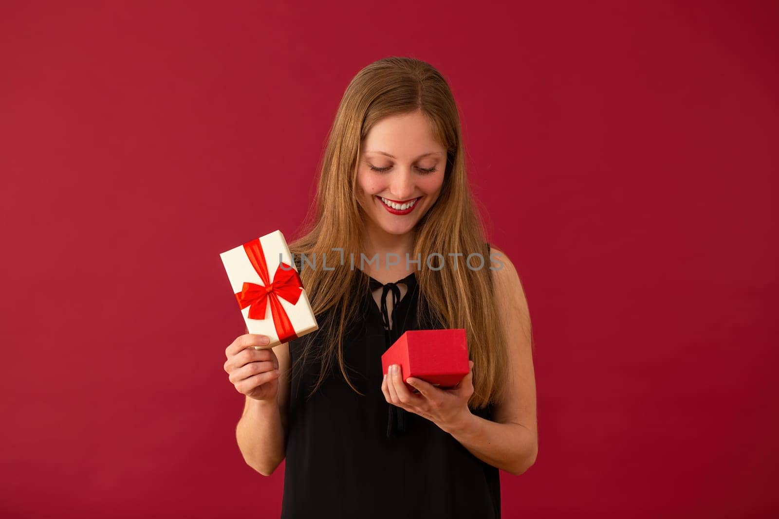 Smiling woman opening the box with ribbon on red background. Happy girl holding valentines present. Blonde female with gift in hand on burgundy backdrop.