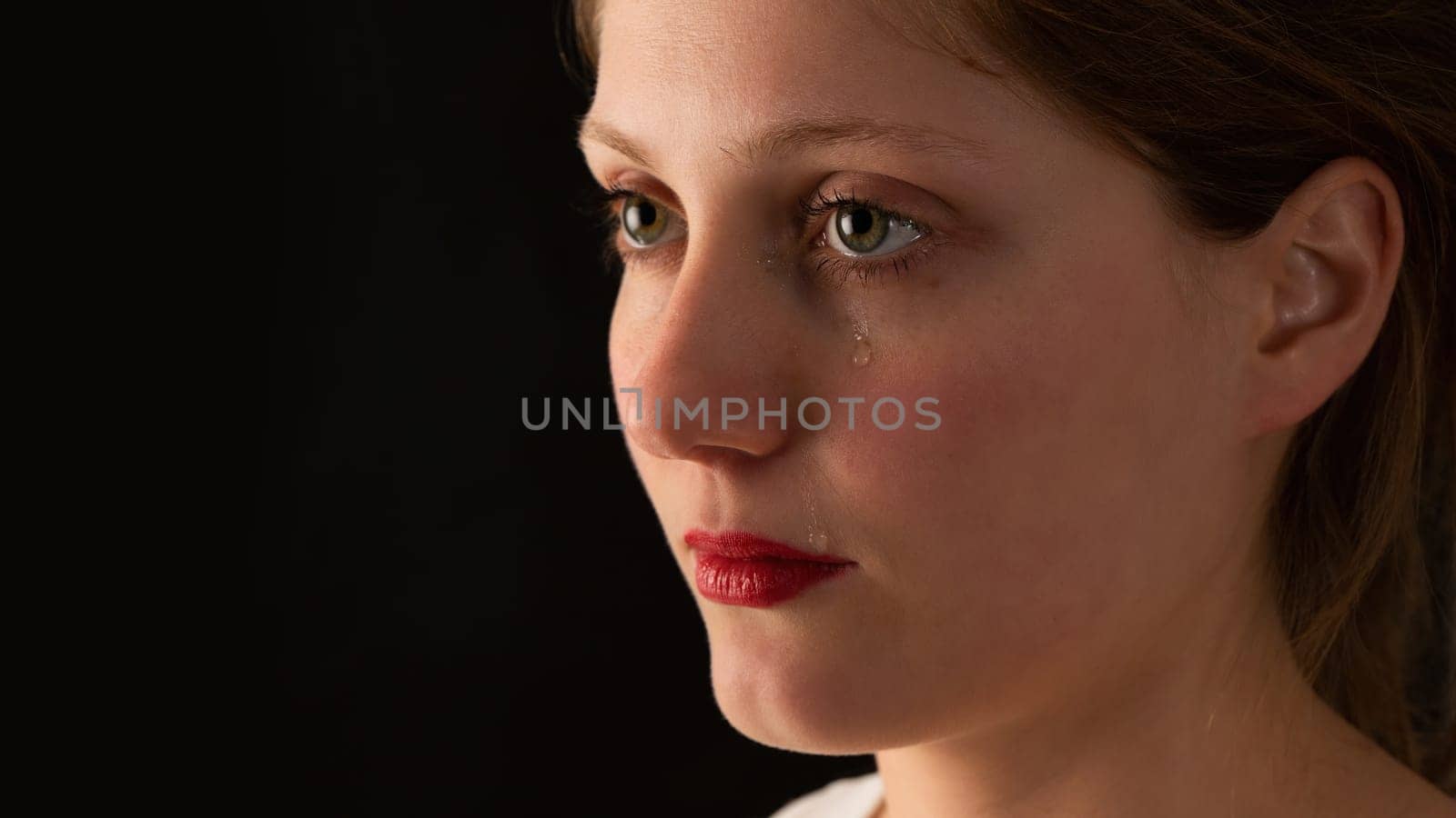 Sad woman crying on black background from close up. Unhappy girl with tears falling from eye on dark backdrop. Younge female feeling upset.