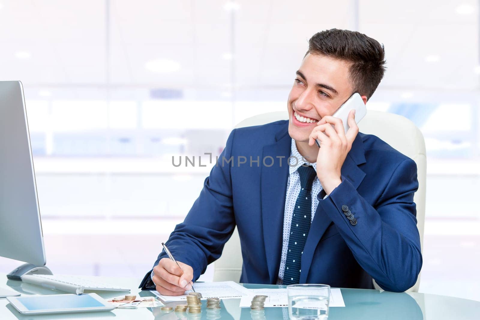 Close up portrait of Attractive young accountant talking on smart phone in office. Young man at desk in front of computer filling out financial documents.