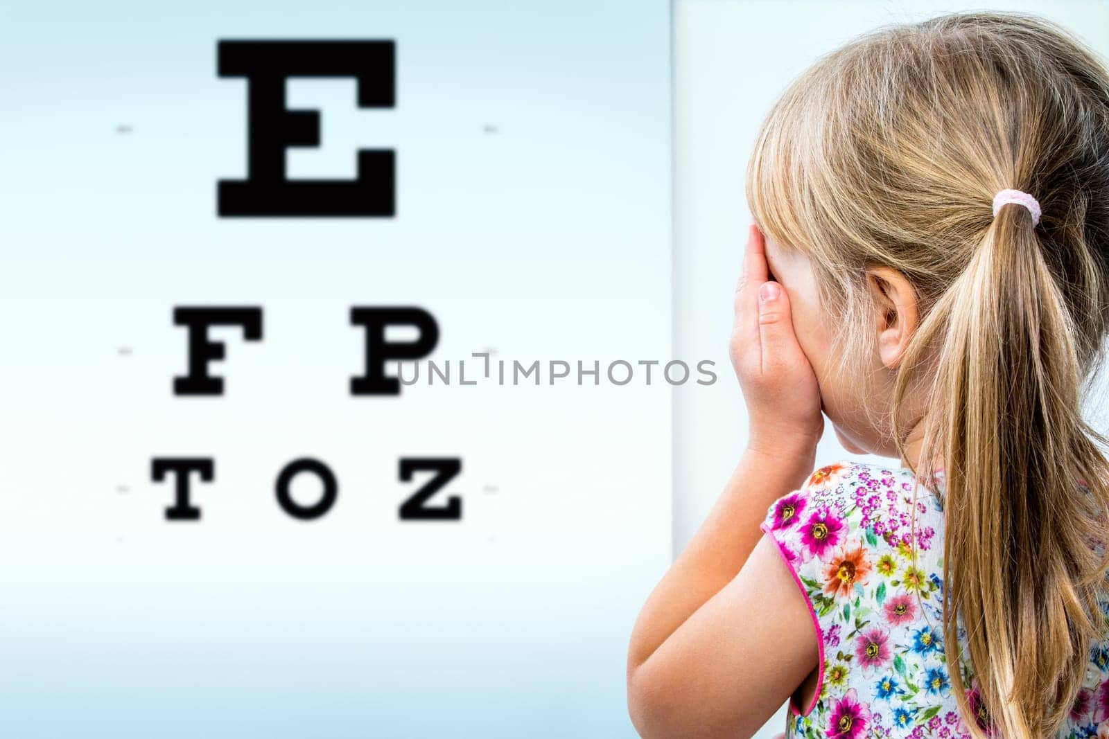 Close up rear view of girl testing eyesight. Infant closing one eye with hand looking at vision test chart.