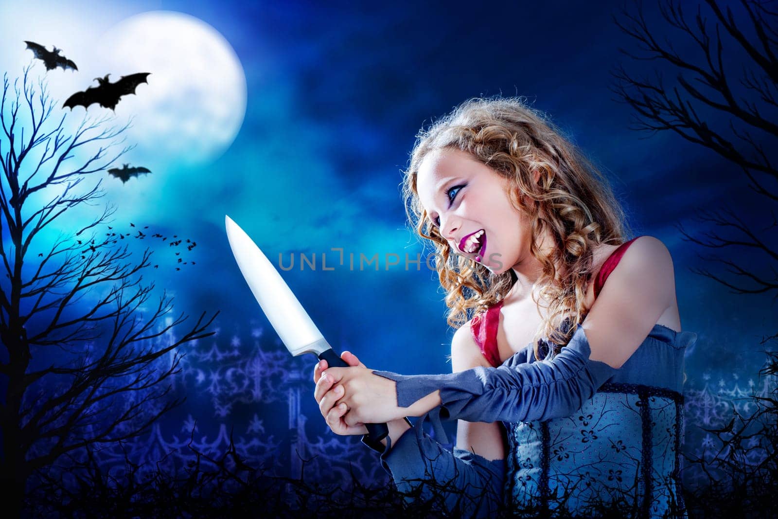Close up horror portrait of Young female vampire holding big knife at dusk.Full moon and flaying bats over graveyard in Background.