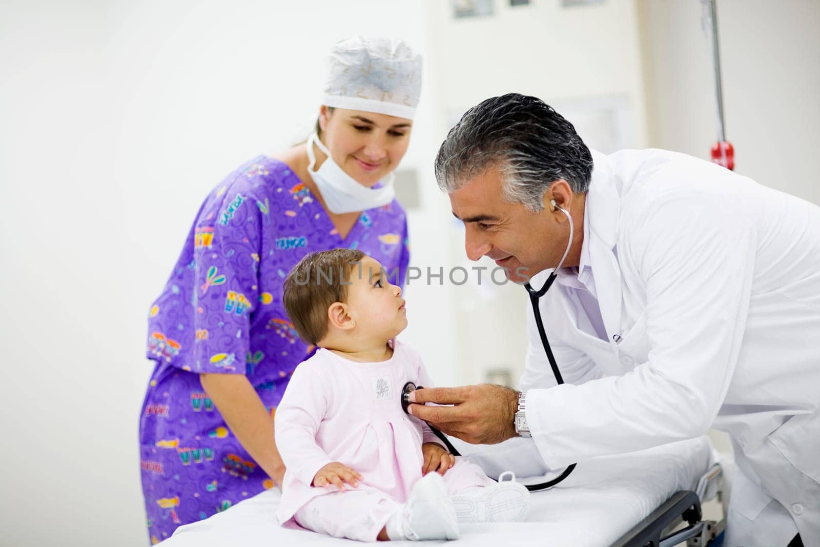 Side profile of a male doctor examining a baby girl with a stethoscope and a female doctor standing near them