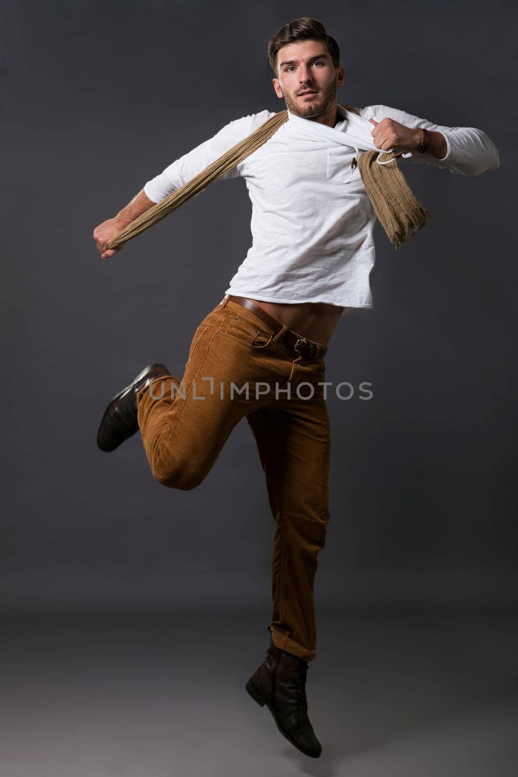 Stylish young man posing in smart casual clothes with a scarf jumps casually in front of the camera against a grey background