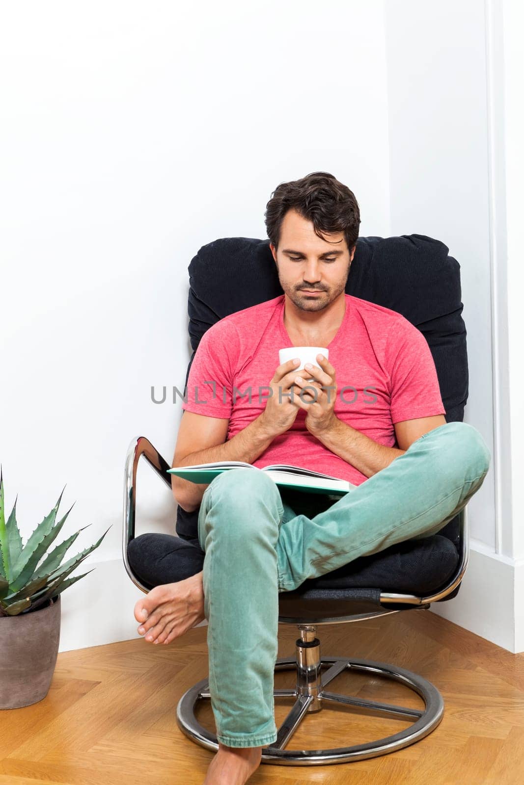 Young man in casual clothes relaxes while reading in an armchair with a drink in his hand