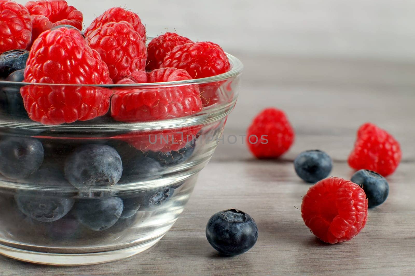 Detail on small glass bowl full of blueberries with raspberries on top, some of the berries spilled on gray wood table.